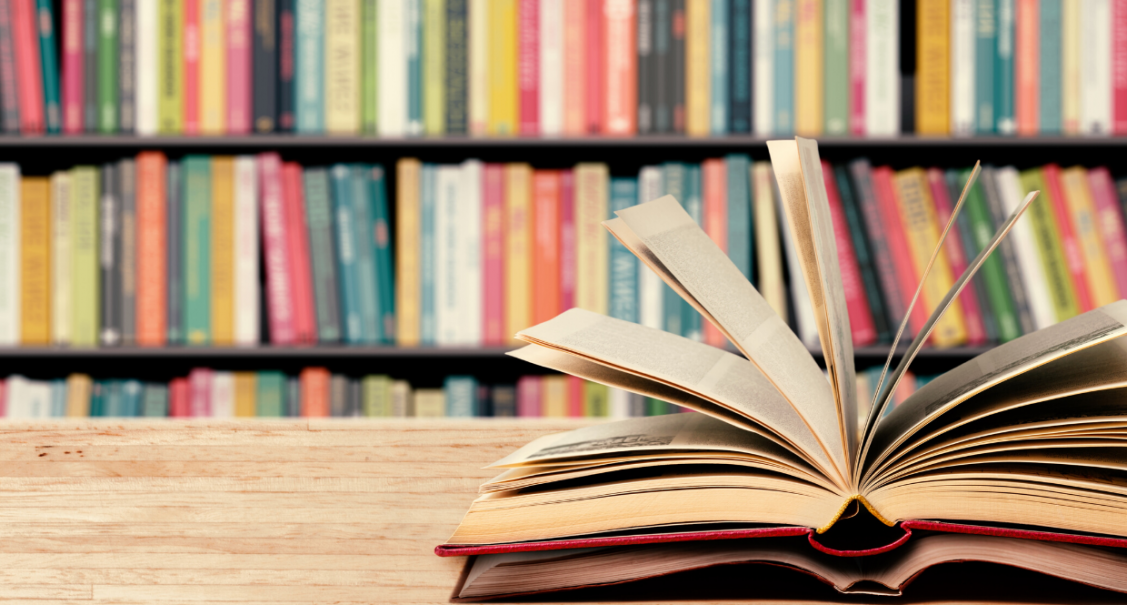 An open book on a table in front of a library shelf of multi-colour books