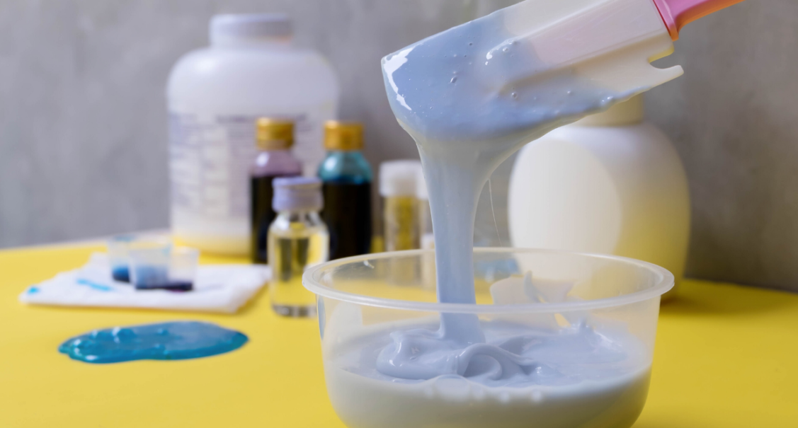 A bowl of cornflour paste dyed purple with food die being stirred by a spatula with food dye and other bottles in the background