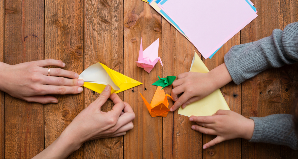 parent and child folding origami birds with brightly coloured paper on a wooden table