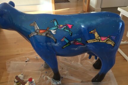 Museum of Cambridge mini-moo, mid-creation by artist Marlis Horgan, showing the night sky painted onto the cow with the rocking horses in full colour.