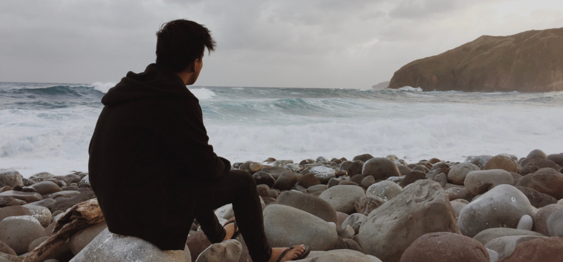 Young man sat on a rock on the shore, looking out to sea on a cloudy day