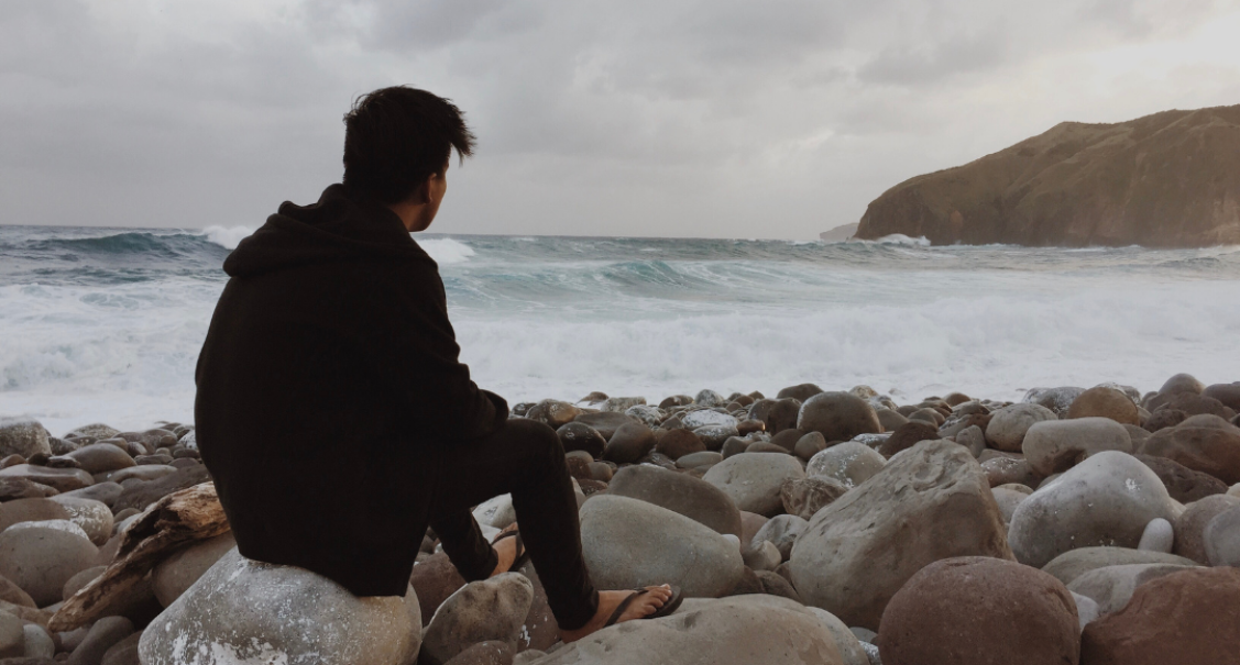 Young man sat on a rock on the shore, looking out to sea on a cloudy day
