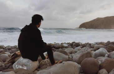 Young man sat on a rock on the shore, looking out to sea on a cloudy day