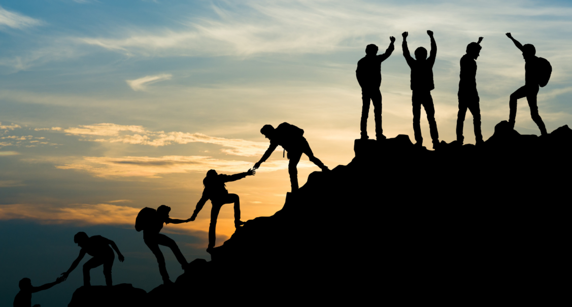 Photo of young people silhouetted against a sunset, helping each other to climb a hill. The photo evokes a feeling of success.