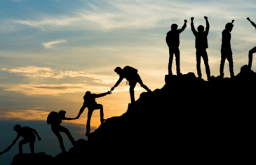 Photo of young people silhouetted against a sunset, helping each other to climb a hill. The photo evokes a feeling of success.